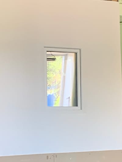 The image shows a white wall with a small, horizontal window in the center. The window frame is light gray. Through the window, a sunlit scene with green foliage and a blue sky is visible. The foliage appears to be trees or bushes. The wall itself is smooth and appears to be drywall. The right edge of the wall shows a slightly different texture and color, possibly indicating the edge of the wall or a transition to another surface. There are small, dark dots visible on this edge, which might be fasteners or imperfections. The lighting suggests it is daytime. No people, text, or specific activities are depicted. The setting appears to be indoors, looking out onto an outdoor scene.