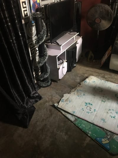 The scene depicts an interior room in Oba-Ile, Nigeria, characterized by dim lighting. In the mid-ground, a black flat-screen television rests on a white, multi-shelf entertainment unit. Below the television, cables are visible on an open shelf. A black speaker unit is positioned to the right of the TV stand. Behind the television setup, the wall is covered with black and white geometric patterned wallpaper, on which a framed picture of two individuals is mounted. To the left of the entertainment unit, a tall, slender object, possibly another speaker or support column, is wrapped with a shaggy grey and white tinsel-like decoration. Further to the left, dark, pleated curtains are drawn.
On the right side of the image, a brown standing fan with a circular grill cover is situated against a reddish-brown wall. Below the fan, a dark clothes iron with a yellow cord is on the floor. A portion of a large white appliance or cabinet is visible in the far right background.
The floor is composed of unpolished concrete. In the foreground, two overlapping rectangular floor mats are present. The top mat is predominantly white with a light blue floral pattern, showing significant wear, soiling, and a torn edge. A second mat, patterned in green and other colors, is partially visible beneath the first. No individuals are visible within the frame.