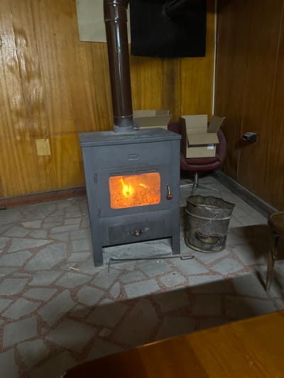 The image displays an interior room located in Sushitsa, Bulgaria. A dark grey, rectangular metal wood-burning stove occupies the central foreground, actively burning with bright orange and yellow flames visible through its glass door. A brown metal chimney pipe extends vertically from the stove's top. Below the main firebox, an ash drawer with a brass-colored knob is present.

To the right of the stove, a dark, dented metal bucket, consistent with an ash container, rests on the floor. A black metal fire poker or tool lies on the floor directly in front of the stove. The floor is covered with irregularly shaped grey flagstone tiles set with reddish-brown grout. The walls are clad in light brown vertical wood paneling.

In the background, to the right of the stove, a burgundy office swivel chair holds two empty, open cardboard boxes. A black electrical outlet is mounted on the wood paneling further to the right. Above the stove, a rectangular black metal plate or vent is affixed to the wall, with visible darkening or soot stains on the surrounding wood paneling. A portion of a brown wooden surface, likely a table, is visible in the lower right foreground. No individuals are present in the scene, and no specific human actions are depicted.