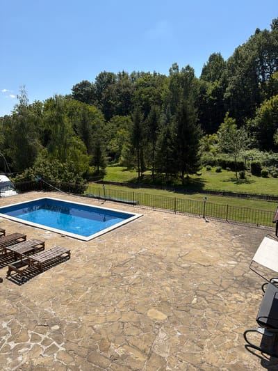 This is an exterior, high-angle, wide shot of a residential property with a swimming pool and surrounding grounds, located in Sokolovo, Bulgaria. The scene is bathed in bright, natural sunlight under a clear blue sky with a few scattered clouds on the horizon. In the foreground, a stone-paved patio area features three wooden lounge chairs arranged parallel to each other. To the left, a portion of a white vehicle is visible.
Dominating the mid-ground is a rectangular swimming pool filled with vibrant blue water, bordered by a white coping. The pool area is further delineated by a dark metal fence that stretches across the property. Beyond the fence, a well-maintained lawn slopes gently upwards, interspersed with various trees and dense green foliage that forms a natural backdrop. The trees, ranging from tall evergreens to deciduous varieties, create a lush, wooded environment. On the far right of the image, a portion of a white outdoor table and chairs is visible, suggesting an area for dining or relaxation. The overall impression is one of a tranquil, secluded outdoor living space.