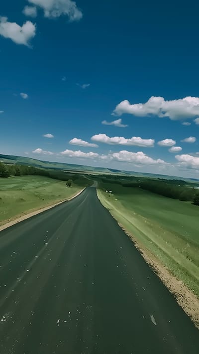 The image depicts an empty, winding asphalt road stretching into the distance, surrounded by lush green rolling hills under a bright blue sky dotted with white clouds. The road appears to be in a rural area, with no vehicles or people visible. The terrain is characterized by open fields and scattered trees, suggesting a natural landscape. The perspective is from a low angle, as if looking from a vehicle traveling on the road. The scene is serene and evokes a sense of open space and tranquility. The specific location in the city of Победа, Russia cannot be confirmed from the visual information provided in the image.