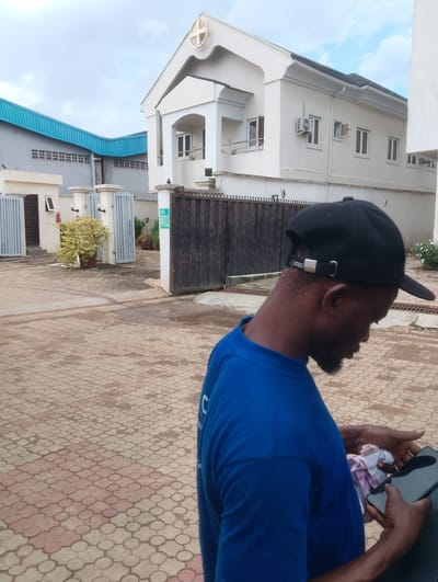 In the city of Ijaiye, Nigeria, a man wearing a blue t-shirt and a black baseball cap is seen from the side, holding a smartphone and a pink and white cloth in his hands. He appears to be looking down at his phone. In the background, a white building with a cross emblem on its facade stands prominently. To the left, another building with a blue metal roof is visible. The ground is paved with interlocking concrete blocks. The scene suggests an outdoor setting, possibly a residential or commercial area, under an overcast sky.