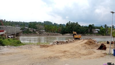 The image depicts a scene near Beunyot, Indonesia, characterized by a construction or repair project along a river. A large, grey metal bridge is visible on the left, spanning the muddy, brown river. The riverbanks are lined with lush green vegetation and palm trees, with a cluster of buildings situated behind the trees.
In the foreground, a construction site is evident. A yellow excavator, branded with "CAT", is positioned on a dirt path. Piles of dirt and large rocks are scattered around, suggesting ongoing earthmoving activities. A detached excavator bucket lies on the ground near the construction area. Two individuals, wearing dark clothing, are seated near a lamppost on the right side of the image, observing the area. The sky is overcast with grey clouds. The overall scene conveys a sense of infrastructure work in a tropical, rural setting.