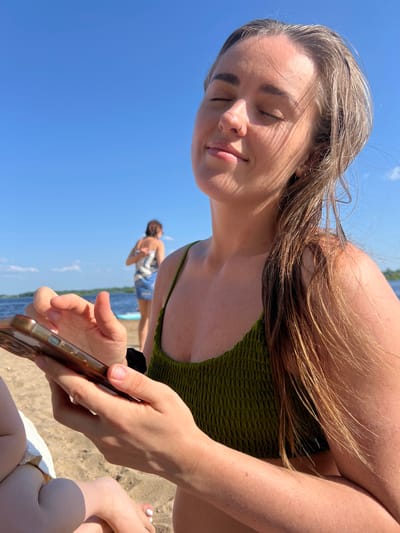 A young woman with blonde hair and closed eyes sits on a sandy beach, holding a smartphone. She wears a green, textured bikini top. Her face is tilted upwards, catching the sunlight. In the background, out of focus, another person stands near the water. The sky is bright blue with a few wispy clouds. The scene appears to be a sunny day at a lake or river.