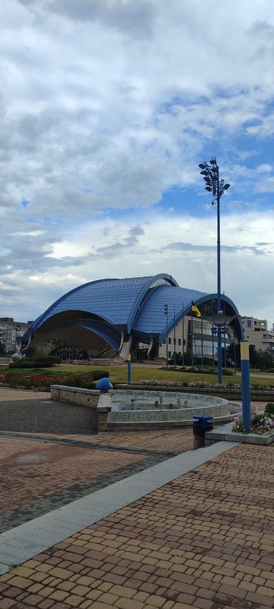 A modern, curvilinear building with a blue, metallic roof dominates the scene, its architecture reminiscent of a shell or wave. The building appears to be a public space, possibly a cultural center or sports arena, with large glass windows reflecting the cloudy sky. In front of the building, a paved plaza features intricate brickwork patterns in shades of brown and gray. A low, circular fountain with a stone border sits in the foreground, its water still. Along the plaza's edge, there are flower beds with pink and red blossoms. A tall, dark blue lamppost with multiple lights stands prominently in the mid-ground, near a Ukrainian flag flying from a pole. The sky is overcast with a mix of gray and white clouds, with patches of blue sky peeking through, suggesting it is daytime but not a bright, sunny day. The overall atmosphere is one of civic architecture and public space, set against a slightly somber sky. There are no people visible in the frame.
