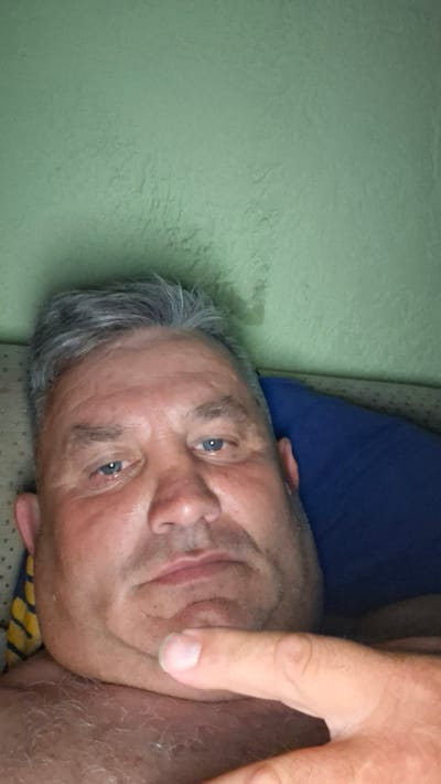 The image is a close-up, selfie-style photograph of an adult male. He has graying hair, light blue eyes, and appears to be lying down. His head is tilted slightly, and his chin is resting on his hand. A blue pillow is visible behind him. The background wall is a light green color. The image is lit from the front, likely by a camera flash. There is no discernible indication of the city of Voinova, Moldova, or any specific urban or rural setting within the image. The focus is solely on the individual's face and upper body.