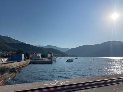 The image depicts a sunlit harbor in Bijela, Montenegro, with a clear blue sky and a bright sun casting a glare on the water. In the foreground, a concrete pier is visible with some metal rails and a purple ribbed cable. The middle ground features a calm, blue body of water dotted with several boats, including a white motorboat with a canopy. Along the left bank, a wall separates the water from a small town with residential buildings, some with red tile roofs, and a large blue structure. A few smaller boats are moored near this bank. In the background, lush green mountains rise, their slopes covered in trees, creating a scenic backdrop. The overall scene conveys a peaceful, coastal atmosphere on a clear day.