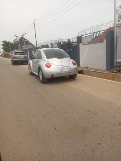 The image shows a street scene in Baccure Kofare, Nigeria, on an overcast day. Two cars are visible on an asphalt road. In the foreground, a silver Volkswagen Beetle is parked. The car has a license plate that reads "MDG 320AE". Behind the Beetle, a green Honda CR-V is also parked, with a license plate that reads "MLA 31UN".

The background features a grey and dark grey wall with a black gate topped with barbed wire. The wall appears to be part of a residential property. Above the wall, a dark grey, pyramid-shaped roof with a water tank is visible. Several utility poles with overhead wires are also present. The sky is a uniform, hazy grey, suggesting cloudy weather. The overall impression is of a quiet residential street. No people are visible in the image.