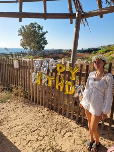 A young woman wearing a white, lace-trimmed tunic and dark gray slides stands on a sandy beach in Palmachim, Israel. She has a white flower crown on her head and a necklace with a small pendant. Behind her, metallic balloons spelling out "HAPPY BIRTHDAY" are attached to a wooden fence. The balloons are gold and silver, with "HAPPY" in silver and "BIRTHDAY" in gold. Beyond the fence, the blue sea is visible, with a tree and vegetation on the coastline. The sky is clear and blue. The scene appears to be outdoors, under a wooden canopy structure, suggesting a celebratory setting.