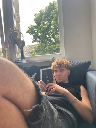 This media file depicts a young man lounging on a couch, holding a smartphone. He is wearing a black t-shirt and dark, distressed shorts. A silver bracelet adorns his left wrist. The man has curly, blonde hair and is looking towards the camera.
The scene appears to be indoors, near a window that looks out onto trees and a residential area, suggesting a suburban or urban setting in Hamm, Germany. Sunlight streams in, indicating it's daytime. A small, black fan is visible on a windowsill, suggesting a warm day. The overall mood is relaxed and casual. The smartphone has a decorative case featuring a cross emblem. There is no visible text.