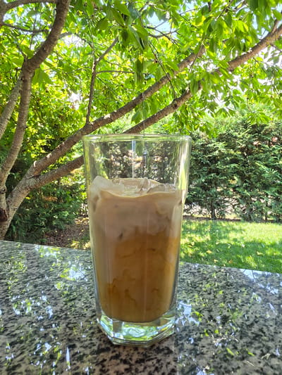 A tall glass of iced coffee sits on a speckled granite table outdoors, likely on a patio. The coffee is a light brown color with a foamy top and visible ice cubes. Behind the glass, lush green trees and bushes form a natural backdrop, with dappled sunlight filtering through the leaves, creating a pleasant, shady atmosphere. The overall scene suggests a relaxed, outdoor setting on a bright day, possibly in the city of Indzhe Voyvoda, Bulgaria, given the context.