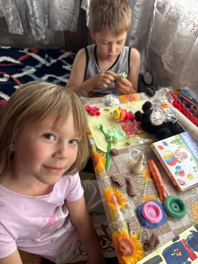 The image shows a young boy and girl engaged in creative play with modeling clay on a table covered with a sunflower-patterned tablecloth. The boy, wearing a gray tank top, is focused on a piece of green clay in his hands, possibly shaping it. In the foreground, the girl, wearing a pink t-shirt with a cartoon character print, looks towards the camera with a slight smile, her eyes wide and attentive. Her blonde hair is neatly styled.

Scattered on the table are various colors of modeling clay, some already molded into shapes like a red figure, a brown piece, and a white cone. A box of "ПЛАСТИЛИН 12" (Plasticine 12) is visible, indicating the type of material being used. Other items include a black stuffed animal, a drawing of a character, and some transparent plastic wrappers. The scene appears to be indoors, with a lace curtain visible on the left, suggesting it might be daytime. The overall mood is one of playful creativity and childhood innocence. The location is identified as Маркидоновка, Russia.