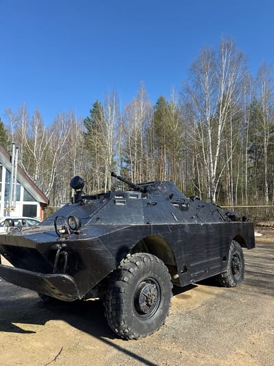 A black, wheeled armored vehicle is parked in an outdoor area, likely in the city of Галево, Russia. The vehicle has large, treaded tires and is situated on a dirt or gravel surface next to a paved area. A building with white-framed windows and a sloping roof is visible in the foreground, partially obscured by the vehicle. In the background, there is a stand of bare trees against a clear blue sky. The scene appears to be taken during daylight hours.