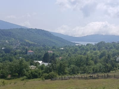 The image depicts a panoramic view of a rural landscape, likely near the city of Nikortsminda, Georgia. In the foreground, a dry, grassy field is bordered by a rustic wooden fence. Beyond the fence, a dense forest of lush green trees covers rolling hills. Scattered amongst the trees are various buildings, including houses with red and white roofs, and a more prominent, light-colored multi-story building with a red roof. In the middle ground, a body of water, possibly a lake or reservoir, is visible in the distance. The background features layered mountain ranges receding into the horizon, with the furthest peaks softened by a hazy sky filled with white, puffy clouds. The overall atmosphere is serene and natural, suggesting a peaceful, undeveloped environment.