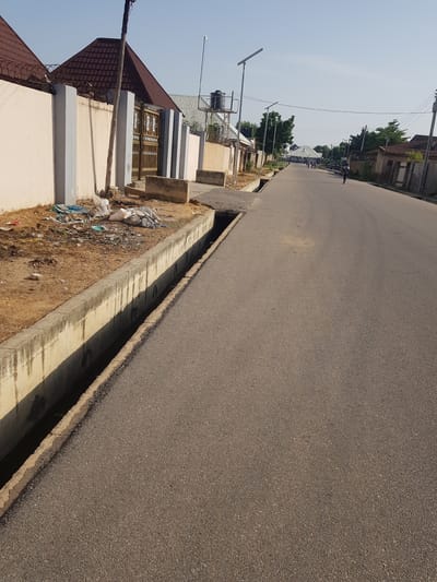 The image depicts a street scene in Palade, Nigeria. The foreground shows a concrete gutter running along the side of an asphalt road. The gutter is partially filled with water and debris. To the left of the gutter is a sandy area with trash piled up. A beige wall with gray pillars runs along the sandy area, topped with barbed wire. A gate with decorative metalwork is visible in the wall. A wooden pole with wires is positioned near the gate.

The road stretches into the distance, with houses and trees lining both sides. Several solar-powered streetlights are visible along the road. In the background, a few people are walking or standing. The sky is clear and blue. The overall impression is of a residential street with some signs of development and potential neglect.