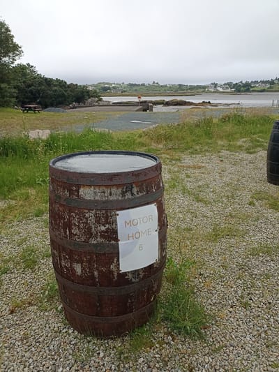 The image shows a wooden barrel with metal bands, standing on a gravel surface in Annagry West, Ireland. A sign attached to the barrel reads "MOTOR HOME 6". In the background, there is a body of water, a green shoreline with trees, and a few buildings. The sky is overcast.