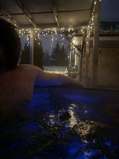 An individual with dark, curly hair is depicted from the upper torso, submerged in blue-lit water within what appears to be an outdoor hot tub or spa. A bundle of dark, slender branches is partially submerged in the water near the individual's right arm. Above the water, a dark-colored metallic canopy or pergola structure is visible, strung with multiple strands of warm white fairy lights. The background reveals an exterior nighttime scene in Базарово, Russia, characterized by snow-covered ground and rooftops. Dark evergreen trees and bare deciduous trees are present in the distance. To the right, a section of a light-colored brick building with a snow-dusted roof is observable.