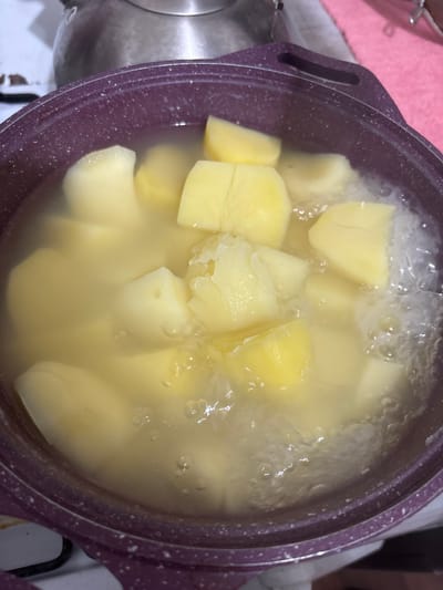The image shows a close-up, overhead view of a purple pot filled with boiling water and chopped potatoes. The potatoes are cut into large, irregular chunks and appear to be cooking. Bubbles rise to the surface of the water, indicating active boiling. The pot is positioned on a stovetop, with a silver kettle visible in the upper left corner and a pink surface to the right. The scene appears to be a domestic kitchen setting. There are no people visible in the image. The location within the city of Княгинино, Russia, cannot be determined from the visual information provided.