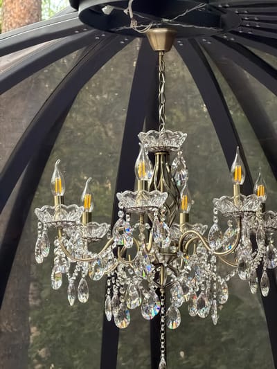 A brass chandelier with crystal accents hangs inside a dark, geodesic dome structure. The chandelier has multiple arms, each supporting a clear, flame-shaped LED bulb and adorned with tiered crystal cups and dangling faceted crystals that reflect rainbow colors. The background is a blurred view of green foliage, suggesting an outdoor setting. The dome's frame is visible as dark, curved beams. The chandelier is the primary subject, positioned centrally and well-lit. The overall impression is one of decorative illumination within a sheltered space. The location is specified as Rakitnitski selski Saveet, Belarus, though this information cannot be visually confirmed from the image itself.