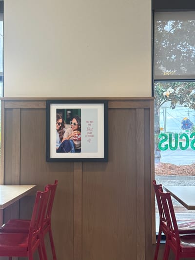The image shows a framed picture hanging on a wooden paneled wall. The picture features two women, both wearing sunglasses, smiling and embracing. Above the picture is a window, through which a view of a building with a sign can be seen. Parts of red chairs are visible on the right side of the image. The picture includes the text "YOU ARE THE Best PART OF TODAY." The scene appears to be indoors. The location is in the city of Stone Mountain, United States of America.