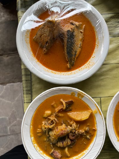 Three white ceramic plates containing reddish-orange curries are depicted from an overhead perspective on a mixed surface in Kutalhoksukon, Indonesia. The uppermost plate holds two substantial pieces of fish, partially submerged in the curry, and is sealed with transparent plastic wrap, showing internal condensation. The central plate, located below and to the left, contains squid and potato chunks in a similar curry, also covered with transparent plastic wrap. A third plate, partially visible on the bottom right, features the same curry and plastic wrap. All plates are white with a subtle gold rim. The surface beneath the plates consists of a yellow-green, textured fabric on the right, juxtaposed with a grey, possibly tiled or concrete, patterned surface on the left. A dark fabric object is partially visible in the lower-left corner of the frame.