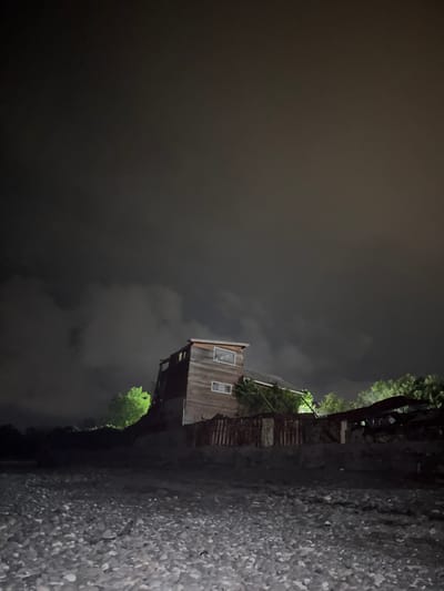 The image depicts a rustic, wooden structure at night in Gumsta, Abkhazia. The building, appearing to be a two-story dwelling or workshop, has visible windows and is constructed with weathered wooden planks. Surrounding the structure is overgrown vegetation, including trees illuminated by an unseen light source, creating a stark contrast against the dark, cloudy sky. The foreground is a rough, rocky terrain, possibly a shoreline or unpaved area. The scene suggests an abandoned or secluded location, with the lighting casting long shadows and highlighting the dilapidated state of the surroundings. No people are visible in the image.