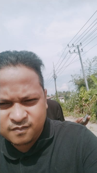 The image is a selfie taken outdoors in Kruengseupeng, Indonesia, featuring a man in the foreground wearing a black collared shirt. He has dark hair and a mustache and goatee. In the background, another person is visible, and a hand is holding a smartphone. The environment includes power lines supported by utility poles, lush green vegetation, and a few buildings in the distance. The sky is bright and overcast.