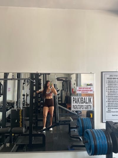 The image shows a woman taking a mirror selfie in a gym located in Cuyo, Philippines. The woman is wearing a brown sports bra and brown shorts, with white sneakers. She is standing in front of a weight rack, holding a smartphone in her right hand. The gym appears to be well-equipped with various weight machines and barbells. There are also signs on the wall, including one in Filipino that says "Pakibalik po ang mga gamit sa tamang lalagyan pagkatapos gamitin. Maraming salamat!", which translates to "Please return items to their proper place after use. Thank you!". Another sign, in English, outlines the gym's rules and regulations. The overall environment suggests a functional and active fitness facility.