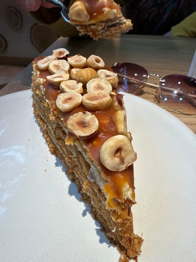 A close-up shot depicts a slice of layered cake topped with what appears to be nuts and a caramel-like glaze on a white plate. To the left of the cake are brown sunglasses. The location is Перепечино, Russia.