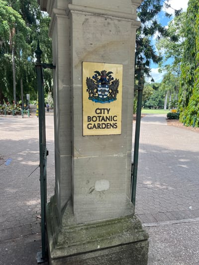 The image captures the main entrance to the City Botanic Gardens in Brisbane, Australia, on a bright, sunny day. The scene is dominated by a substantial stone gatepost on the left side, to which a rectangular, yellowish sign is affixed. This sign proudly displays the name "CITY BOTANIC GARDENS" in dark, vertical capital letters on its right. To the left of the text, an intricate crest or coat of arms is embossed in dark colours with highlights of blue and white, featuring heraldic elements such as a shield, supporting figures, and a decorative top element.

A sturdy green metal fence or railing extends horizontally from the stone pillar, forming a clear boundary for the entrance. Beyond this barrier, paved pathways, featuring both hexagonal paver patterns and smoother asphalt, lead into the expansive gardens. The background is filled with the lush, dense foliage of numerous trees, indicating a rich botanical environment. Sunlight filters through the canopy, casting distinct shadows across the pathways. In the far distance along one of the garden paths, a small, indistinct figure of a person can be observed, suggesting the typical use of the gardens by visitors. The clear sky and strong illumination point to a pleasant daytime setting.