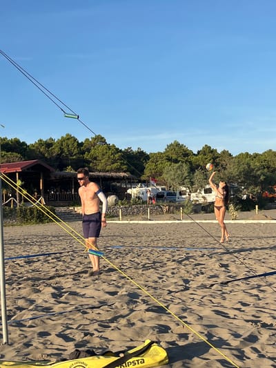 In Pistula, Montenegro, two individuals are playing beach volleyball on a sandy court under a clear blue sky. A man, wearing sunglasses and dark blue shorts, stands on the left side of the net, facing the right. He has a light-colored arm sleeve on his left arm and appears to be in mid-game. On the right side of the net, a woman in a brown bikini is in the process of serving a volleyball, her arm extended upwards. The beach court is bordered by a net, with yellow and blue lines visible on the sand. In the background, a line of green trees borders the sandy area. Beyond the trees, several recreational vehicles and a building with a thatched roof are visible, suggesting a campground or resort area. The overall scene depicts a recreational activity taking place in a coastal environment.