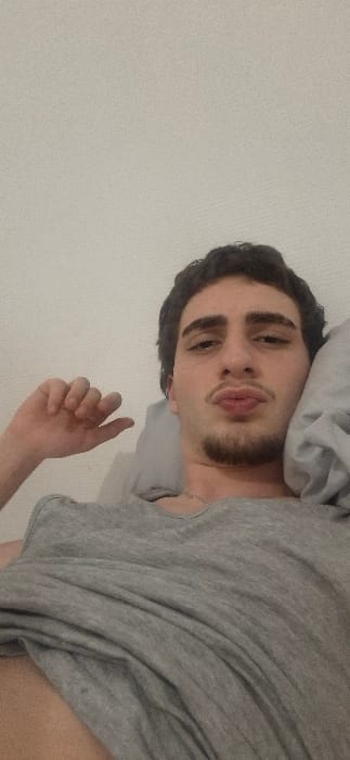A young man with dark hair and a short beard lies on his side in bed. He wears a gray t-shirt and rests his head on a gray pillow. His face is turned towards the camera, and his lips are slightly pursed in a kiss. One hand is visible, extended with fingers loosely curled. A white wall is visible in the background. The image is rotated slightly.