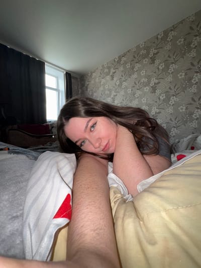 The image depicts a young woman lying on a bed in what appears to be a bedroom. She has long, dark brown hair and is looking directly at the camera with a relaxed expression. Her eyes are green. She is wearing a grey t-shirt. The bed is covered with light-colored bedding, including a white and grey striped pillowcase with a red heart design and a yellow blanket. In the background, there is a window with dark curtains, and the wall is covered in floral wallpaper. The lighting suggests it is daytime. This scene is located in the city of Башкултаево, Russia.