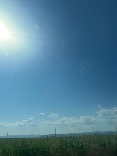 The image captures a wide-angle view of a rural landscape under a bright, clear blue sky. In the upper left quadrant, the sun is visible, emitting strong rays and creating a lens flare effect. The sky transitions to a lighter blue towards the horizon, dotted with scattered white clouds.

The lower half of the image is dominated by a vast, green field, likely agricultural, with tall grasses and crops. Two utility poles are visible in the field, spaced apart. In the background, a range of low, rolling hills or mountains stretches across the horizon, their shapes softened by distance and haze. The overall scene suggests a sunny day in a rural setting, possibly in the city of Lemnia, Romania, given the context. There are no people or animals discernible in the image.