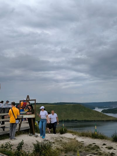 The image depicts an outdoor scenic overlook in Horaivka, Ukraine, under an overcast sky. In the foreground, a dirt path leads down from an elevated wooden viewing platform situated on the left. On the platform, several individuals are visible, including a man in a yellow short-sleeved shirt facing right and a woman in a dark top standing beside a wooden, framed structure. In the center-right foreground, a woman wearing a white t-shirt, blue jeans, and a white baseball cap walks towards the viewer, holding a white smartphone. Behind her, a man in a white polo shirt and dark pants also walks along the path. Two green and yellow striped markers are positioned on the right side of the path. The mid-ground reveals a wide, dark body of water, likely a river, winding through a landscape of green, undulating hills and distant plains. A prominent, rounded hill rises directly across the water in the mid-ground. The upper half of the frame is dominated by a solid layer of grey clouds.