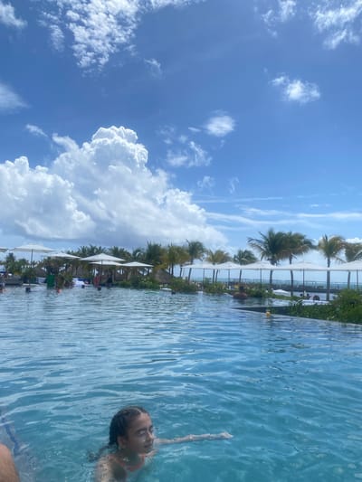 Here's a detailed description of the image:

**Location:** Marina Maroma, Mexico. The image appears to be taken at a resort featuring an infinity pool.

**Environment:** The sky is bright blue with large cumulus clouds. There are palm trees and white sun umbrellas lining the edge of the pool. The pool water is clear and light blue.

**People:** A person is swimming in the pool in the bottom right corner of the image. The person appears to be female. The pool edge is lined with sun umbrellas and beach chairs where several people are seated.

**Objects:** The main objects are an infinity pool, umbrellas, palm trees, chairs, and clouds.