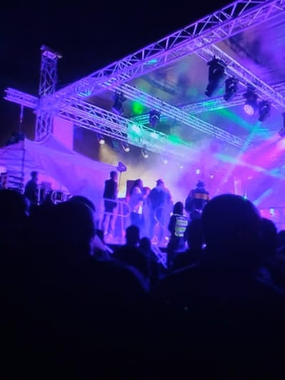 This image depicts an outdoor night concert or event taking place at a venue in Dodoma, Tanzania. The scene is illuminated primarily by stage lighting in hues of purple and blue, interspersed with green and magenta beams cutting through a visible atmospheric haze.
In the foreground, a dense crowd of spectators is present, their heads and upper bodies appearing as dark, indistinguishable silhouettes, indicating a low-light environment from the audience perspective.
The midground showcases a raised stage structure. On the stage, approximately 6-8 individuals are visible. One person on the far left appears to be operating a sound or lighting console behind a table. A second individual is positioned on a small elevated platform slightly right of center. A figure on the right side of the stage is clearly identifiable by a bright yellow or neon green reflective vest, characteristic of event staff or security personnel. Other individuals on the stage are less defined but appear to be actively engaged in the performance or event operations.
The background is dominated by a substantial metallic truss structure overhead, forming the stage's support system. Multiple professional stage lights, including moving head fixtures, are mounted on this truss, projecting intense, colored light beams through the hazy air. A large, light-colored screen or backdrop is partially visible behind the stage. The night sky is dark above the stage structure.