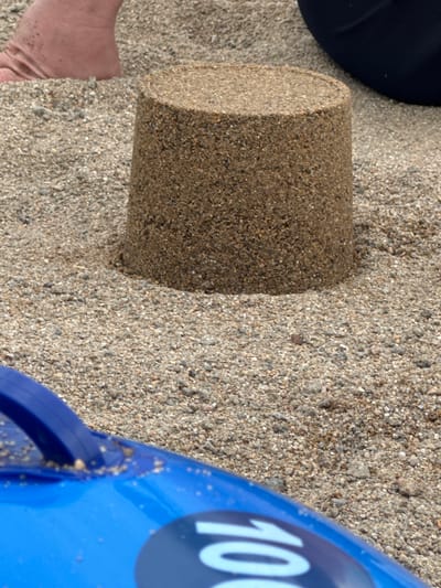 A close-up, high-angle shot shows a cylindrical sand mold pressed into grainy sand. To the left of the mold, a portion of a person's left foot and ankle are visible, with the skin appearing smooth and lightly tanned, and a few grains of sand adhering to it. The sand is a mixture of light brown and grey pebbles and grains. To the right of the sand mold, a bright blue object, possibly a bucket or toy, is partially visible. It has a smooth, curved surface and a large white "100" printed on it in a bold font. The edges of the blue object appear slightly worn and have some sand particles clinging to them. The scene suggests an outdoor setting, likely a beach or sandy area, with a focus on objects used for sand play. The lighting suggests it is daytime, possibly sunny given the clarity of the image. The image is cropped tightly around the subjects and objects, leaving no background context other than the sand.