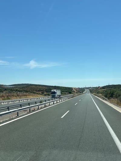 The image shows a highway with a clear blue sky above. The road has multiple lanes and is lined with metal guardrails. On the left side of the road, there is a hill with dry vegetation and some solar panels. A white truck is driving on the highway. In the distance, the road curves into the distance, surrounded by rolling hills covered in green and brown vegetation. There are some power lines visible on the hills in the far distance. The scene is one of a highway cutting through a rural landscape on a sunny day. The image was taken from a vehicle moving along the highway.