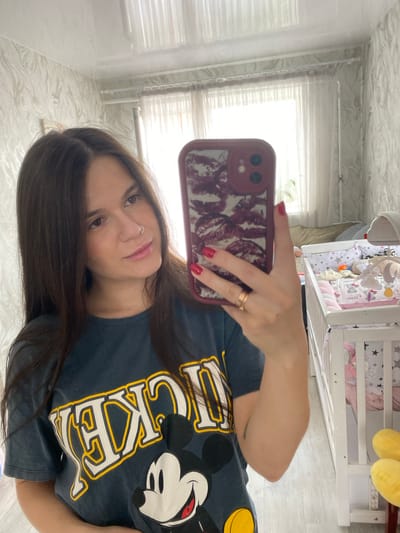 A young woman with long, dark hair and a nose ring takes a selfie in a room. She is wearing a dark grey t-shirt with a graphic of Mickey Mouse and text. Her right hand, with red nail polish, is holding a phone with a red case. She is wearing a gold ring on her left ring finger. In the background, there is a white crib filled with bedding and stuffed animals. The room has patterned wallpaper and sheer curtains. The location is identified as Romny, Ukraine.