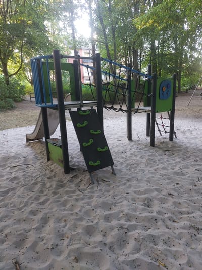 The image shows an outdoor playground area with a sand surface. A climbing structure is the central focus, featuring a blue railing, green panels, a slide, a climbing wall with green grips, and a rope net. The playground is situated amidst trees, with a building visible in the background. The setting appears to be a park or residential area in Herzogenrath, Germany. There are no people or animals present in the image. The sand is textured with footprints and scattered leaves and twigs. The sun is shining through the trees, casting dappled light on the scene.