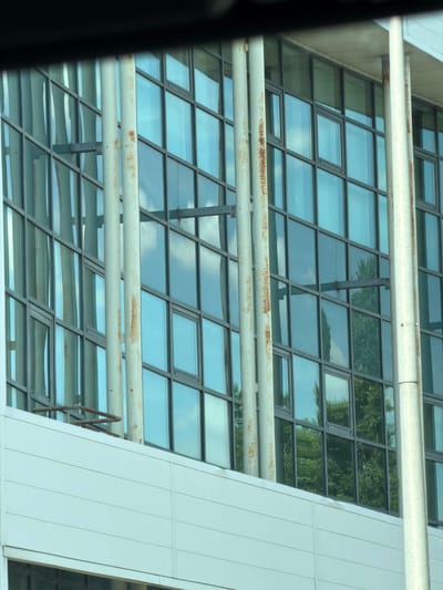 The image shows the exterior of a modern building with a large glass facade. The facade is composed of numerous rectangular panes arranged in a grid pattern, reflecting the sky and surrounding trees. Two horizontal pipes, appearing weathered with some rust, run across the lower portion of the building, partially obscuring the glass. The building's architecture suggests a contemporary design, possibly an office building or public institution.
The time of day appears to be daytime, with bright daylight illuminating the scene and casting reflections. The presence of clear blue sky and some clouds in the reflections indicates fair weather. The image seems to be taken from a slightly angled perspective, possibly from another building or a vehicle, looking up at the structure. The architectural style and the reflections of greenery suggest an urban or suburban setting in a location with trees, such as Évry-Courcouronnes, France. No people are visible in the frame. The focus is on the building's structure and its interaction with the environment through reflections. There is no visible text in the image.