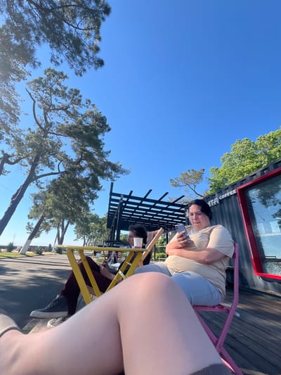 A person is sitting at a table outside a coffee shop. The person is holding a phone in their hand and looking at it. The coffee shop appears to be made out of a shipping container, with a red trim around a window. There is a tree with green leaves behind the coffee shop. The sky is blue, indicating a clear day. The sun is shining, casting shadows on the wooden deck where the table and chairs are located.

The person is wearing a light-colored t-shirt and light-colored pants. They have dark hair. The table is yellow, and the chairs are pink and yellow. A black fence is behind the table.

The image appears to be taken in the daytime, likely late morning or early afternoon, given the bright sunlight and clear sky. The setting appears to be an outdoor seating area for a coffee shop. There is visible text on the side of the shipping container that reads "COFFEE". The overall mood is relaxed and casual, with the person engrossed in their phone.