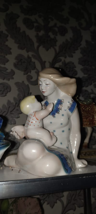 This is a close-up, indoor photograph of a porcelain figurine depicting a mother and child. The mother, with fair skin and blonde hair swept to the side, is seated and cradling a baby. She is wearing a white dress adorned with blue polka dots and a blue neckline trim featuring delicate floral patterns in blue and gold. Her eyes are closed, and her expression appears serene. The baby, with fair skin and blonde hair, is propped up on the mother's lap, facing the mother. The baby is wearing a white garment with small red polka dots. The figurine is placed on a shelf against a backdrop of wallpaper with a dark, ornate damask pattern. To the right of the figurine, a portion of another decorative object, possibly a gilded box or stand, is visible. The lighting in the image is dim, suggesting an indoor setting, likely a home or display cabinet. There is no visible text in the image. The context of Baghdad, Uzbekistan is provided, but there are no visual cues in the image that directly suggest this specific location.