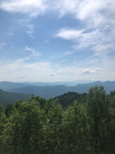 This is a landscape photograph of a mountain range under a partly cloudy blue sky. In the foreground, a dense forest of green trees is visible. The middle ground consists of rolling hills covered in trees, and further in the distance, layered mountain ranges fade into a hazy blue. The sky is dominated by a bright blue expanse with scattered white clouds, some of which are wispy and translucent. The overall impression is one of natural beauty and vastness. The image is likely taken from an elevated viewpoint, offering a panoramic view of the mountainous terrain. The location is Abaza, Russia, a city situated in the southern part of the Republic of Khakassia, known for its mountainous landscapes and proximity to the Sayan Mountains.