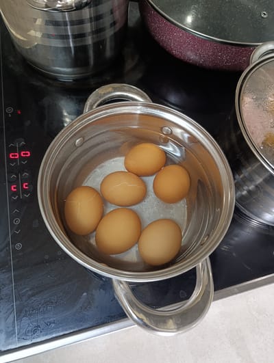 A close-up, overhead shot shows six brown eggs in a pot of water on a black electric stovetop. The eggs appear to be in the process of being boiled, as evidenced by the water and the placement on the stove. Several of the eggs have visible cracks in their shells. To the left of the pot, a digital display shows red illuminated numbers and symbols, indicating the stovetop's controls. The stovetop itself shows some smudges and water marks. In the background, the edges of two other pots or pans are visible. The pot with the eggs is made of stainless steel and has two handles. The lighting is overhead and appears to be from an indoor source. The image does not provide any contextual information to determine its location in Šabac, Serbia.