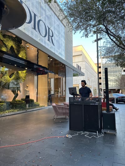 A DJ stands behind a black DJ booth with a laptop set up, in front of a Dior store on a city sidewalk. A sound system is positioned to the DJ's right. A long, orange extension cord runs along the sidewalk. The Dior store has large windows with mannequins and displays inside. A tree overhangs the sidewalk, and other buildings are visible in the background.