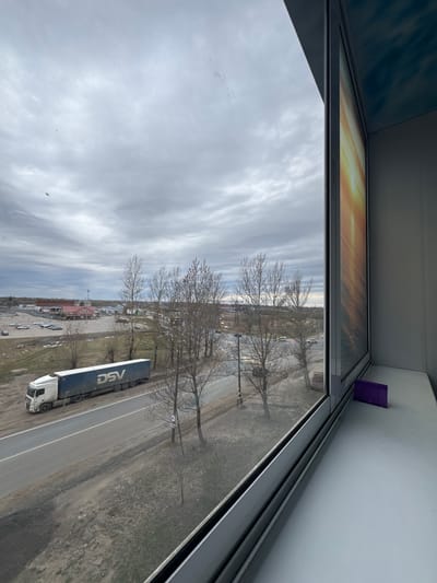 View from a window, possibly an upper story of a building, showing a street scene in Kladovitsy, Russia. Overcast sky with grey clouds. Bare trees lining the road. A large DSV-branded truck is driving on the road. Parking lot with several cars. Purple object visible on the inside window ledge.
