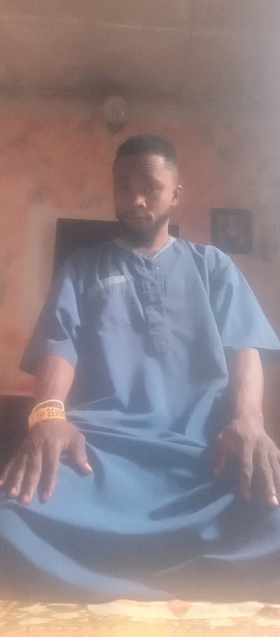 A medium shot depicts a man in a light blue tunic-style shirt. He has short, dark hair and a beard. A yellow bracelet is visible on his left wrist. His hands are resting in his lap. In the background, a blurred television and framed photo can be seen against a pale orange wall. A ceiling light fixture is visible near the top of the frame. The image appears slightly hazy.