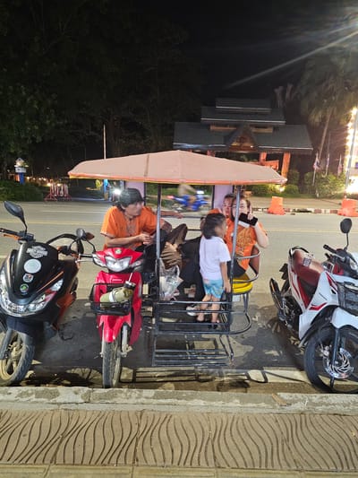 This media file depicts a nighttime scene in Ban Klong Haeng, Thailand, featuring a motorcycle sidecar, its passengers, and surrounding elements.

The main subjects are the people riding in the sidecar. Two adults, possibly parents, are seated, with one woman holding a phone and looking at it. A young child is also in the sidecar, appearing to rest or sleep. The sidecar itself is attached to a red motorcycle. Another white and red motorcycle is parked nearby, positioned at an angle to the sidecar.

The setting appears to be a street at night. Streetlights and lights from nearby buildings illuminate the scene, casting shadows. The ground is paved with textured tiles. There are some trees and foliage visible, suggesting an outdoor environment. Orange traffic cones are present along the side of the road.

The activity seems to be a family or group of people traveling or waiting, possibly as part of a local transportation service common in some parts of Thailand. The overall mood is calm, with no overt signs of distress or excitement.

Notable details include the time of day being nighttime, indicated by the artificial lighting and the dark sky. The presence of Thai flags in the background hints at the location. On the black motorcycle, there are stickers, one of which appears to be a "Harley-Davidson" logo, and another with what looks like a cannabis leaf. A water bottle is attached to the red motorcycle.

There is no explicit weather information, but the presence of people in casual clothing and the general scene suggest it's likely a warm evening.